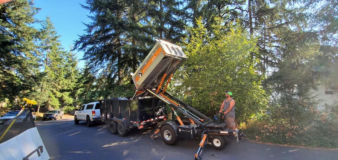 Dump trailer unloading debris in a residential area surrounded by tall trees.