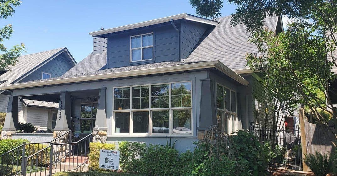 Gray two-story house with large windows and green shrubs, for sale sign in front yard.
