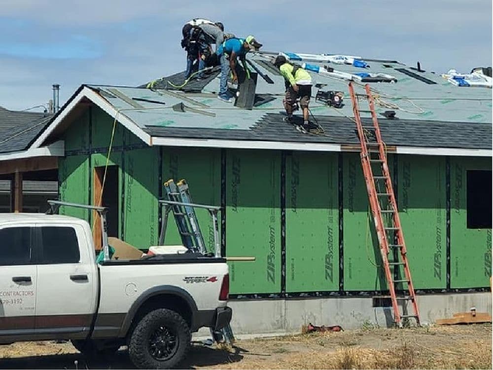 Construction workers installing a roof on a house, with tools and a truck nearby.