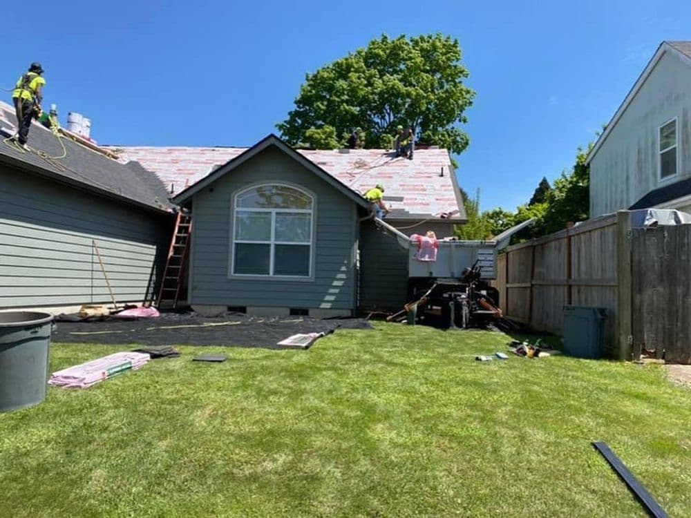 Roofers installing shingles on a house under a clear blue sky with green grass and trees.