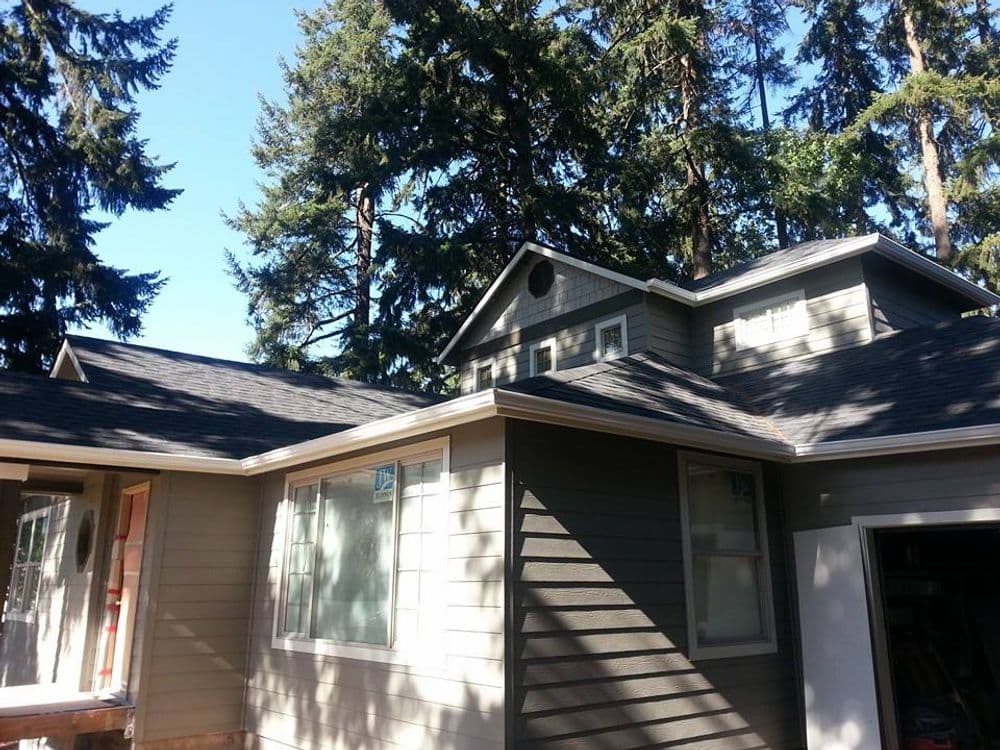 Modern house exterior with new roofing, surrounded by tall pine trees and clear blue sky.