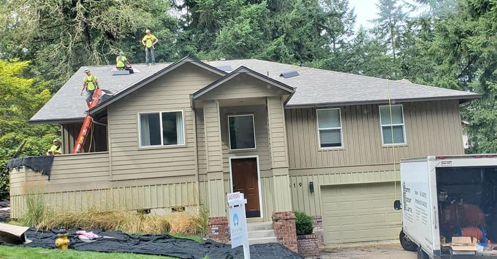 Roofing crew working on a suburban home in a wooded area, truck parked nearby.